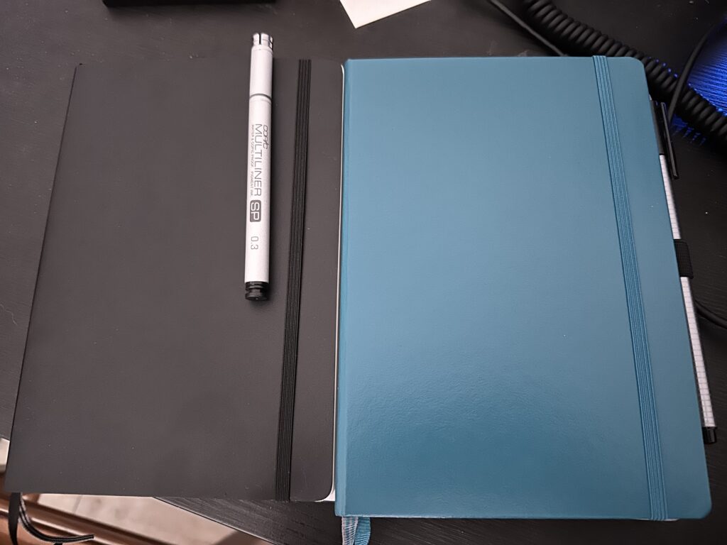 A black and a green notebook, side by side, each with a pen attached.